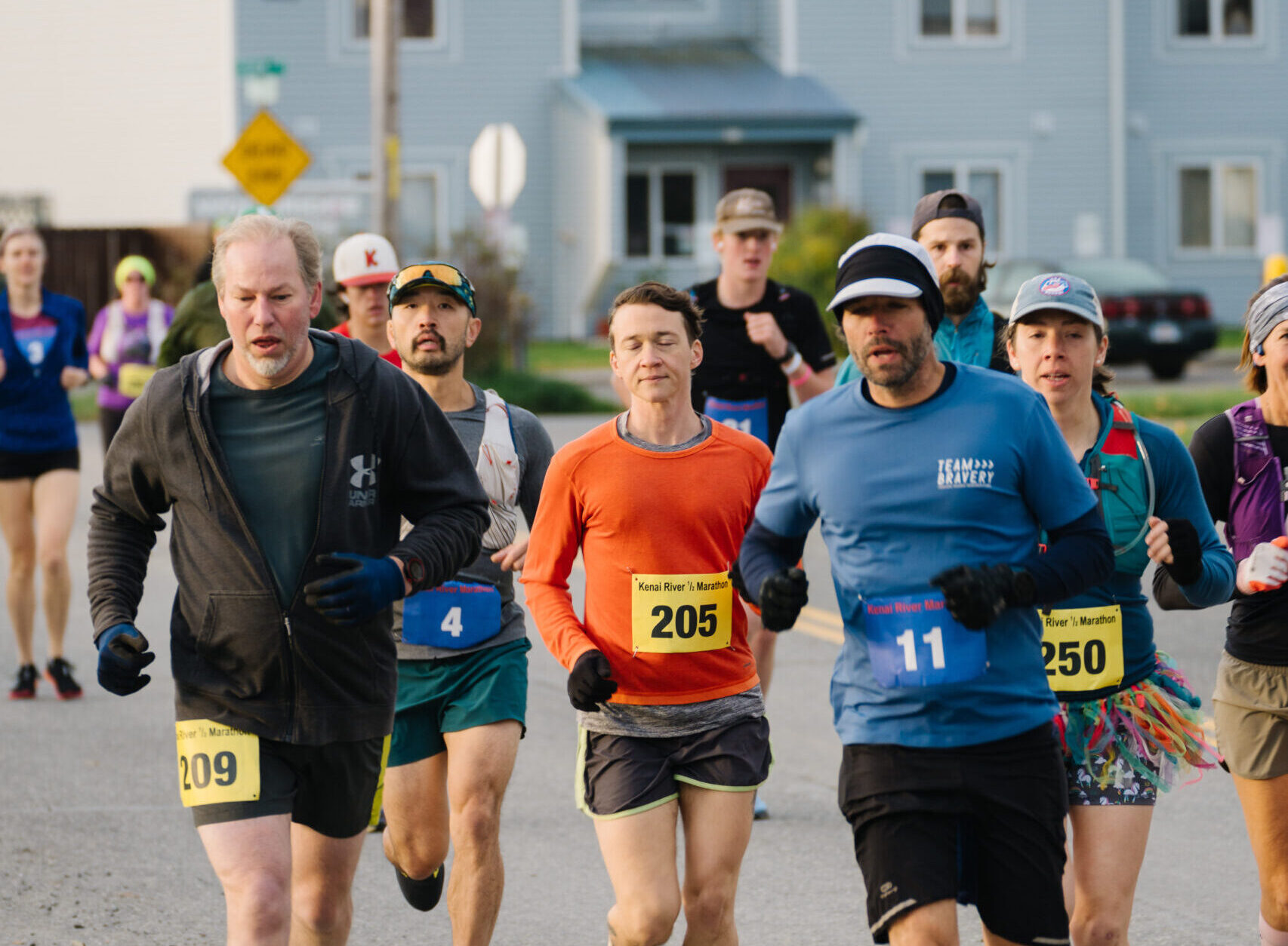 A group of marathon runners in mid-stride on a paved road, participating in the Kenai River Marathon. The runners are dressed in cool-weather gear, including gloves and long-sleeved shirts. The runner in the foreground wears an orange shirt and is focused on the race, while others are seen close behind. Residential houses are visible in the background under a clear sky.